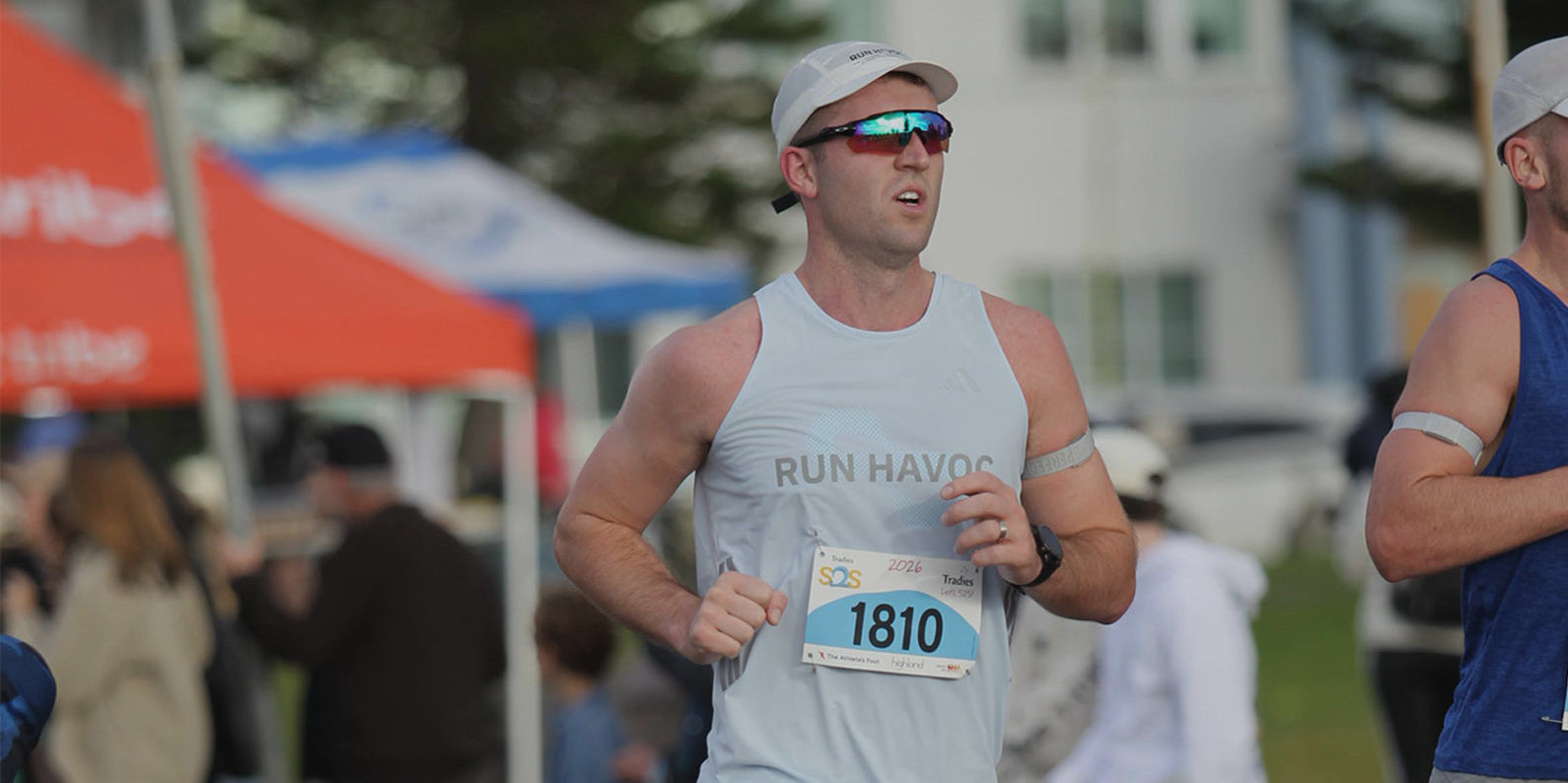 A male runner wearing a white tank top with 'RUN HAVOC' and the number 1810 on his bib, running during an outdoor race with tents and spectators in the background.