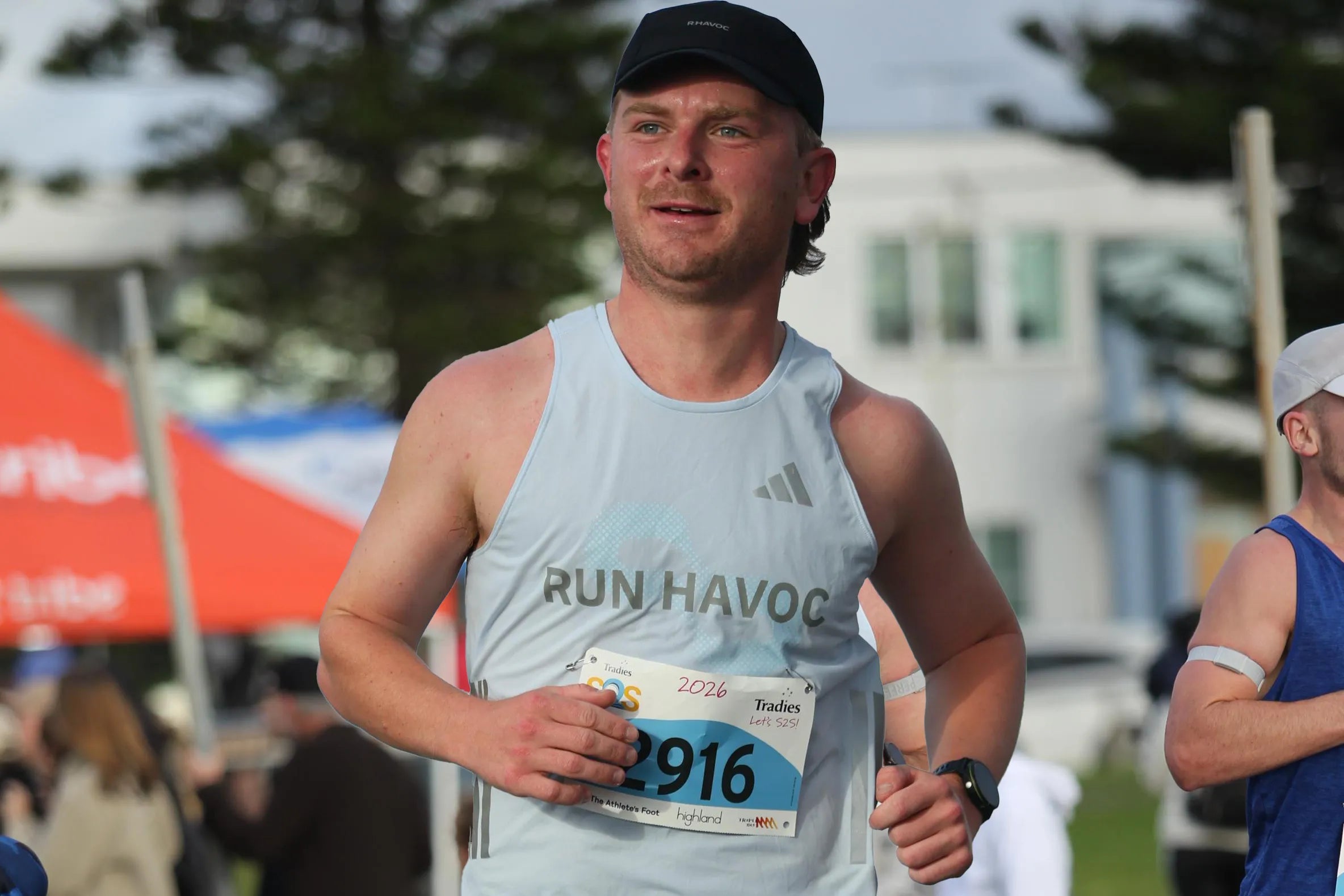 A male runner wearing a black cap, a light blue tank top with 'RUN HAVOC' and an Adidas logo, and a race bib number 2916, captured mid-stride outdoors at a race.
